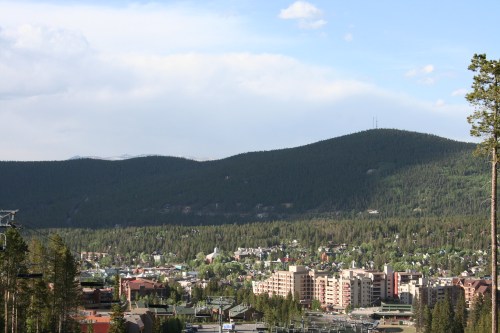 A scenic view of a mountain town surrounded by lush forests and distant mountains under a clear blue sky.