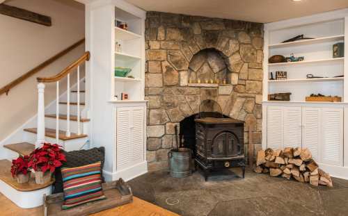 Cozy living space featuring a stone fireplace, wooden shelves, a staircase, and a stack of firewood.
