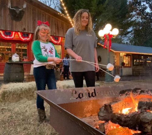 Two girls roast marshmallows over a fire pit at a festive outdoor setting, decorated for the holidays.