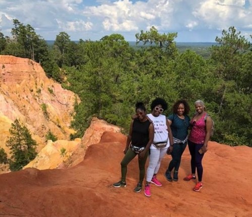 Four women stand on a rocky ledge with a scenic view of trees and hills in the background.