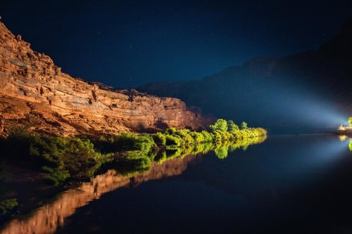 A serene night scene of a river reflecting rocky cliffs and trees, illuminated by a distant light under a starry sky.