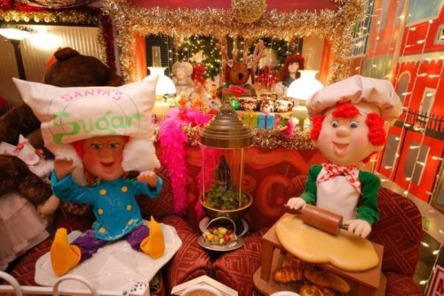 Two festive, oversized characters in a decorated room, one with a pillow hat and the other baking, surrounded by holiday decor.