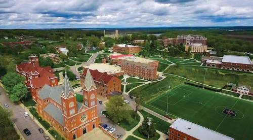 Aerial view of a college campus featuring buildings, green spaces, and a sports field under a cloudy sky.