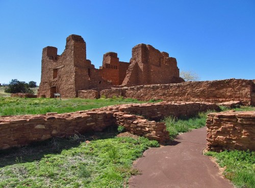 Ruins of an ancient stone structure surrounded by green grass and a clear blue sky. Pathway leads through the site.