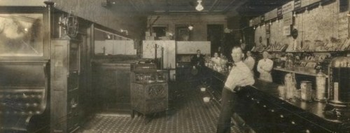A vintage interior of a shop with a counter, shelves, and people working, showcasing early 20th-century decor.