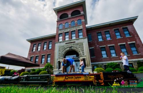 A man adjusts a model train on tracks in front of a historic brick building, with children observing nearby.