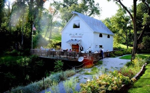 A white barn-style building with a deck, surrounded by greenery and people enjoying the outdoor space.