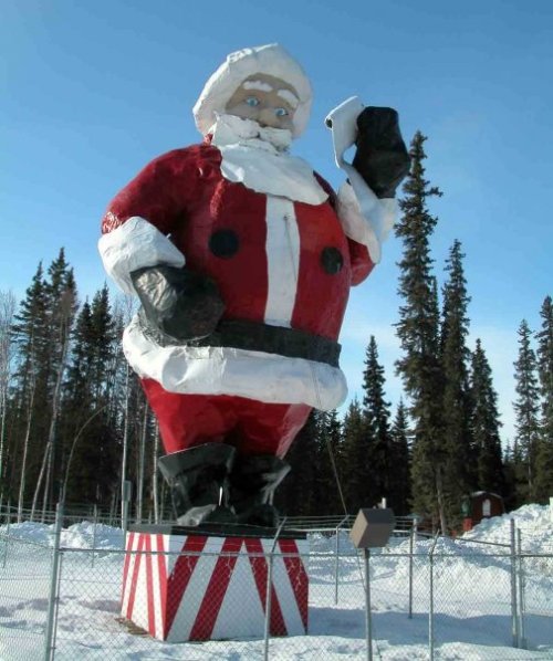 A giant Santa Claus statue holding a phone, surrounded by snow and trees, with a fence in the foreground.