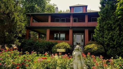 A two-story house with a porch, surrounded by colorful flowers and a statue in the foreground.