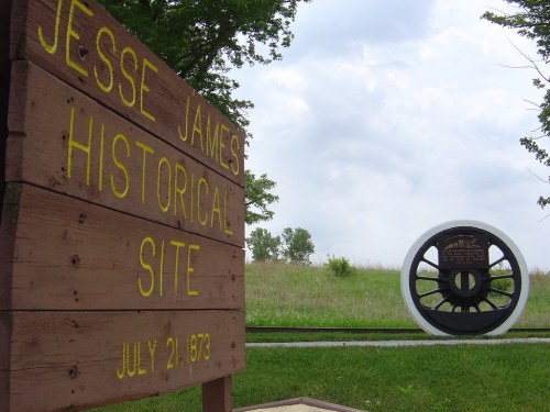 Sign for Jesse James Historical Site with a wheel monument in the background, set against a cloudy sky.