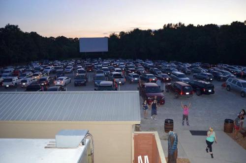 A crowded drive-in movie theater at dusk, with numerous cars parked and a large screen in the background.