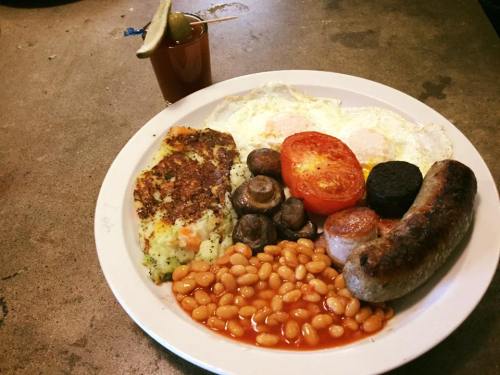A hearty breakfast plate with eggs, sausage, mushrooms, baked beans, tomato, and a side of a cocktail.
