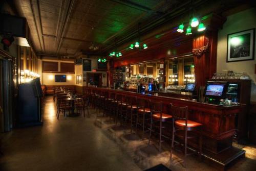 A dimly lit bar with green pendant lights, wooden counter, and empty tables, creating a cozy atmosphere.