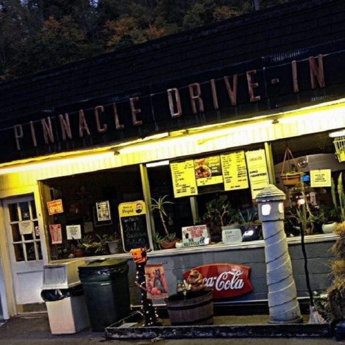 A vintage drive-in restaurant with a neon sign, menu board, and outdoor seating, surrounded by greenery.