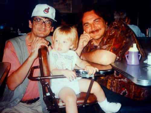 Two men and a toddler sit together at a table, smiling. The toddler is in a high chair, wearing a light shirt.