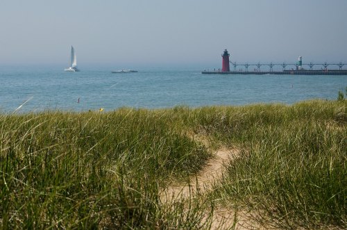 A sandy path leads through tall grass to a calm lake with a red lighthouse and sailboats in the distance.