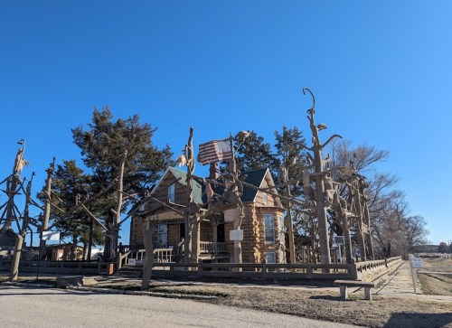 A unique log cabin surrounded by wooden sculptures and trees under a clear blue sky.