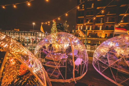 Cozy outdoor dining igloos illuminated with string lights, set against a city skyline and holiday decorations.