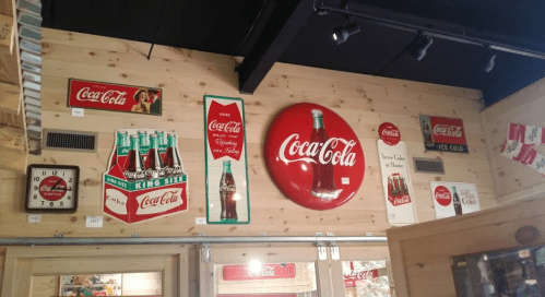 Vintage Coca-Cola signs and memorabilia displayed on a wooden wall, featuring bottles and a large round logo.