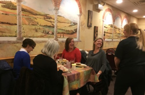 A group of four women laughing at a table in a cozy restaurant, with a server attending to them. Colorful wall murals in the background.