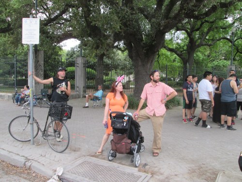 A man on a bike stands by a sign, while a couple with a stroller looks on, surrounded by a crowd in a park.