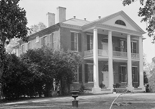Historic brick mansion with large white columns, surrounded by greenery and a spacious lawn.