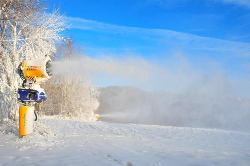 A snowmaking machine sprays artificial snow in a winter landscape with trees and a clear blue sky.