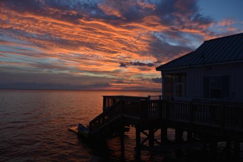 A vibrant sunset paints the sky in orange and purple hues over calm waters, with a wooden dock and house in silhouette.