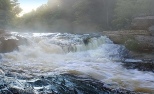 A misty waterfall cascades over rocky terrain, surrounded by lush greenery and soft morning light.
