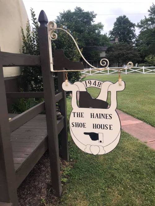 Sign for "The Haines Shoe House" established in 1948, hanging from a decorative bracket near a grassy area.