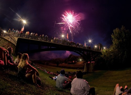 Crowd watching fireworks from a bridge, with people sitting on the grass below, under a night sky.