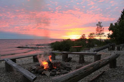 A serene lakeside scene at sunset, featuring a campfire surrounded by logs and colorful clouds in the sky.