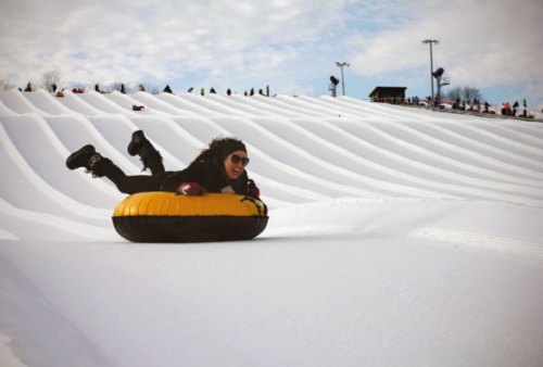 A person in sunglasses joyfully sleds down a snowy hill on a yellow inflatable tube.