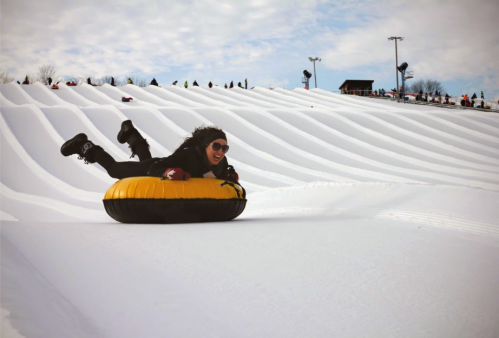 A person in sunglasses joyfully sleds down a snowy hill on a yellow and black tube.