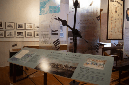 Exhibit display in a museum featuring historical panels, a large clock, and photographs of Natchez.