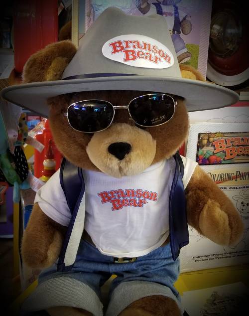 A plush bear wearing sunglasses, a gray hat, and a shirt with "Branson Bear" printed on it, dressed in denim overalls.