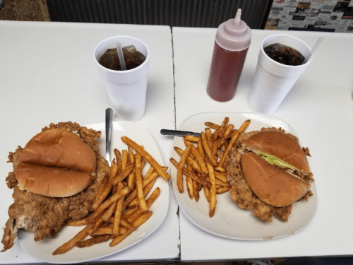Two plates with fried chicken sandwiches, fries, and two cups of soda on a table.