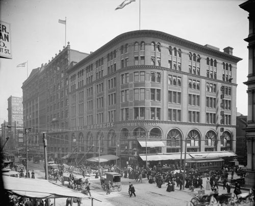 Historic street scene featuring a large building with "Sibel Brothers" signage, bustling with people and horse-drawn carriages.