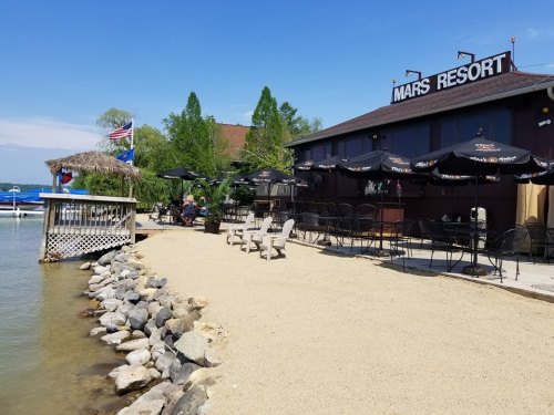 A lakeside resort with outdoor seating, umbrellas, and a sandy shore under a clear blue sky.