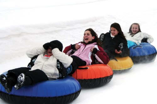Four girls are laughing and tubing down a snowy hill, wearing winter jackets and enjoying a fun day in the snow.
