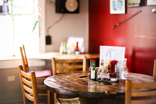 A rustic wooden table in a cozy restaurant, featuring condiments and a menu, with warm red walls in the background.
