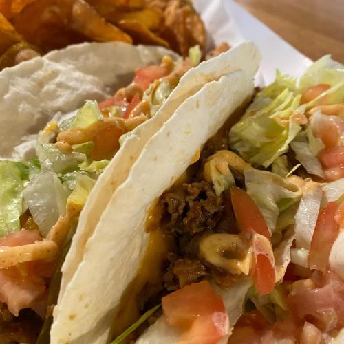 Two soft tacos filled with ground beef, lettuce, and diced tomatoes, served with a side of crispy sweet potato chips.
