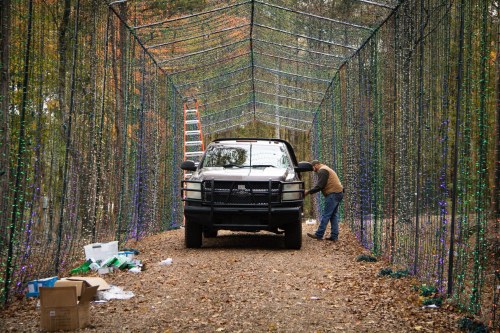 A pickup truck is parked on a gravel path under a canopy of colorful lights, while a person adjusts decorations nearby.