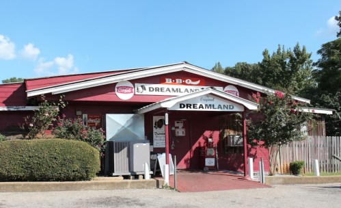 Exterior of Dreamland BBQ restaurant, featuring a red building, signage, and a clear blue sky.