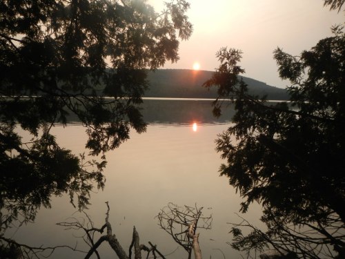 Sunset over a calm lake, framed by trees, with reflections on the water and a hazy mountain in the background.