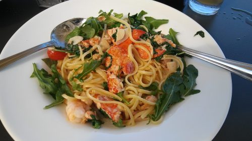 A plate of pasta with lobster, cherry tomatoes, spinach, and herbs, garnished with two serving spoons.