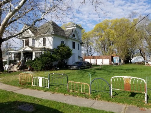 A large, white Victorian house with a tower, surrounded by green grass and old bed frames in the foreground.
