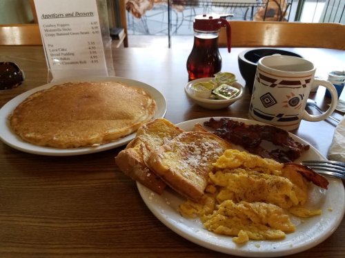 A breakfast plate with scrambled eggs, bacon, French toast, and a side of pancakes, with syrup and coffee on the table.