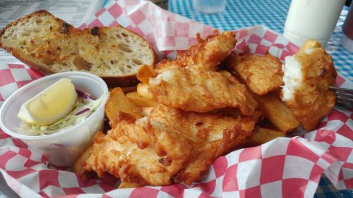A plate of crispy fried fish fillets with fries, a slice of bread, coleslaw, and a lemon wedge in a red-checkered basket.