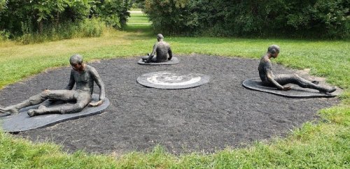 Three seated sculptures on a circular patch of black gravel, surrounded by grass and trees.